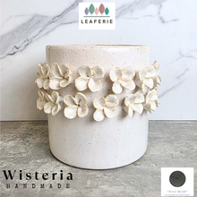 Load image into Gallery viewer, A handmade ceramic planter pot with a textured off-white glaze and a band of raised white plumeria flowers around the middle, set against a marble-patterned background with brand text overlays.