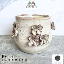 Load image into Gallery viewer, A handmade ceramic planter pot with a textured beige glaze and raised floral details, shown on a gray stone surface against a white marble background. Brand logos for LEAFERIE and Evania are displayed on the image.