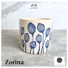 Load image into Gallery viewer, An off-white ceramic planter with a blue hand-painted pattern of stylized flowers, displayed on a gray surface against a white marble background. The brand LEAFERIE is visible at the top, and the name 'Zorina' is shown at the bottom left.