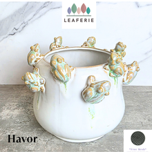 Load image into Gallery viewer, A white ceramic planter pot decorated with multiple 3D frog sculptures in a light green and gold glaze, set on a gray stone surface against a white marble background. The brand logo 'LEAFERIE' is visible at the top, and the text 'Havor' is at the bottom left.