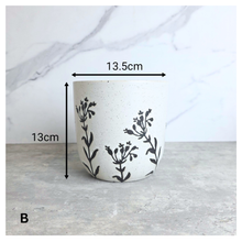 Load image into Gallery viewer, A white speckled ceramic planter with black botanical leaf patterns, shown with dimension arrows indicating a width of 13.5cm and a height of 13cm on a gray countertop against a white marble background.