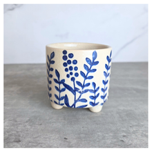 Load image into Gallery viewer, A small ceramic planter pot with a cream-colored glaze and blue painted botanical leaf and berry patterns, sitting on a gray surface against a light gray background.