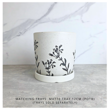 Load image into Gallery viewer, A speckled off-white ceramic planter with a black botanical leaf pattern, shown with a matching white saucer on a gray stone surface against a white marble background. Text at the bottom reads 'MATCHING TRAYS MATTE TRAY 12CM (POTB) (TRAYS SOLD SEPARATELY)'.