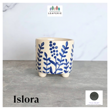 Load image into Gallery viewer, A small ceramic planter pot with a cream-colored glaze and a blue botanical leaf pattern, standing on three small feet. The image features a white marble background, a gray stone surface, the brand name 'LEAFERIE' at the top, the product name 'Islora' at the bottom left, and a mesh filter indicator at the bottom right.
