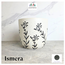 Load image into Gallery viewer, An off-white ceramic planter with black botanical leaf patterns and black speckles, displayed on a gray stone surface against a white marble background. The brand logo 'LEAFERIE' is visible at the top, and the name 'Ismera' is written at the bottom left.
