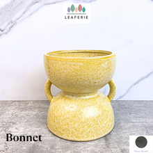 Load image into Gallery viewer, A yellow speckled ceramic planter pot with two side handles, shown on a gray stone surface against a white marble-patterned background. The brand name LEAFERIE is visible at the top, and the text 'Bonnet' is visible at the bottom left.
