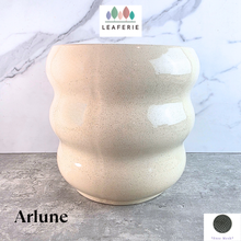 Load image into Gallery viewer, A ceramic planter pot with a wavy, rounded silhouette and a speckled cream-beige glaze, labeled 'Arlune' by the brand LEAFERIE, set against a gray marble-patterned background.
