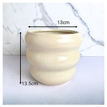 Load image into Gallery viewer, A ceramic planter pot with a wavy, ribbed design in a speckled cream-beige glaze, set against a white marble-patterned background. The brand name 'LEAPERIE' is visible at the top, and the product name 'Petelle' is shown at the bottom left.
