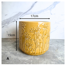 Load image into Gallery viewer, A yellow glazed ceramic planter with embossed wildflower patterns, displayed on a gray stone surface against a white marble background. The brand LEAPERIE is visible at the top, and the product name 'Avara' is shown at the bottom left.
