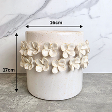 Load image into Gallery viewer, A cylindrical ceramic planter pot with a speckled off-white glaze and a band of raised textured flowers around the middle. Black dimension arrows show a width of 16cm and a height of 17cm against a marble-patterned background.
