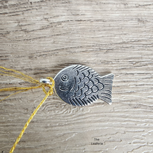 Load image into Gallery viewer, Silver fish-shaped pendant on a yellow thread against a wooden background
