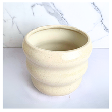 Load image into Gallery viewer, A ceramic planter pot with a wavy, ribbed design in a speckled cream-beige glaze, set against a white marble-patterned background. The brand name 'LEAPERIE' is visible at the top, and the product name 'Petelle' is shown at the bottom left.
