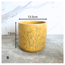 Load image into Gallery viewer, A yellow glazed ceramic planter with embossed wildflower patterns, displayed on a gray stone surface against a white marble background. The brand LEAPERIE is visible at the top, and the product name 'Avara' is shown at the bottom left.
