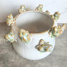 Load image into Gallery viewer, A ceramic planter pot with a glossy white glaze, featuring multiple raised frog motifs in a light green and ochre-brown glaze around the exterior rim and sides.

