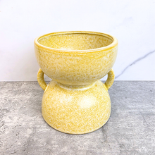 Load image into Gallery viewer, A yellow speckled ceramic planter pot with a wide top bowl and two side handles, sitting on a gray textured surface against a white wall.
