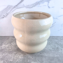 Load image into Gallery viewer, A ceramic planter pot with a rounded, tiered design in a speckled cream-beige glaze, set against a gray concrete surface and a white marble-patterned background.
