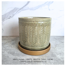 Load image into Gallery viewer, A ceramic planter pot with embossed leaf patterns in a speckled sage green glaze, sitting on a matching round matte brown tray. The background is a gray stone surface with a white marble-patterned wall behind it.
