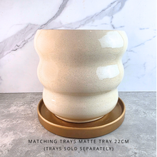 Load image into Gallery viewer, A matte ceramic planter pot with a wavy, rounded silhouette in a warm cream-beige speckled glaze, shown with a matching round tray. The text overlay at the bottom reads 'MATCHING TRAYS MATTE TRAY 22CM (TRAYS SOLD SEPARATELY)'.
