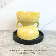 Load image into Gallery viewer, A yellow speckled ceramic double-bowl planter with side handles, displayed on a round black tray. The image is set on a gray stone surface with a white marble-patterned background, and includes a text overlay at the bottom stating 'MATCHING TRAYS MATTE TRAY 15CM (TRAYS SOLD SEPARATELY)'.
