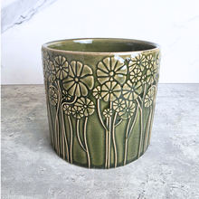 Load image into Gallery viewer, A cylindrical ceramic planter with a glossy olive green glaze and raised cream-colored floral patterns, set against a light gray concrete surface and white marble background.
