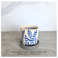 Load image into Gallery viewer, A small round ceramic cup or candle holder with a blue floral pattern on a cream base, shown with a matching dark gray saucer. The text at the bottom reads 'MATCHING TRAYS - MATTE TRAY 9CM (TRAYS SOLD SEPARATELY)'.
