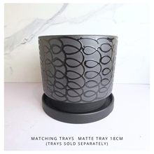 Load image into Gallery viewer, A matte dark gray cylindrical planter with a black interlocking oval pattern, shown with its matching saucer. The background is a white marble-patterned surface with a white floor, and text at the bottom reads 'MATCHING TRAYS MATTE TRAY 18CM (TRAYS SOLD SEPARATELY)'.
