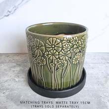 Load image into Gallery viewer, An olive green glazed ceramic planter with embossed daisy-like floral patterns, shown on a round black saucer. The planter is set on a gray stone surface against a white marble-patterned background, with text at the bottom stating 'MATCHING TRAYS - MATTE TRAY 15CM (TRAYS SOLD SEPARATELY)'.
